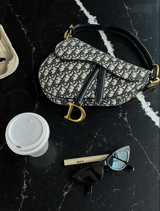 Dior saddle bag oblique monogram gold hardware on a black marble table with black cat eye sunglasses a cup of coffee and refy brow product